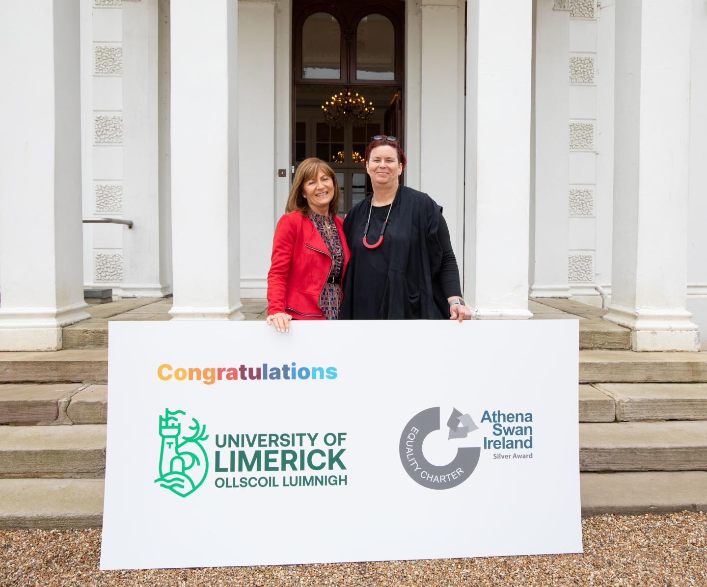 TWo women hold a sign with University of Limerick and Athena Swan logo outside impressive building