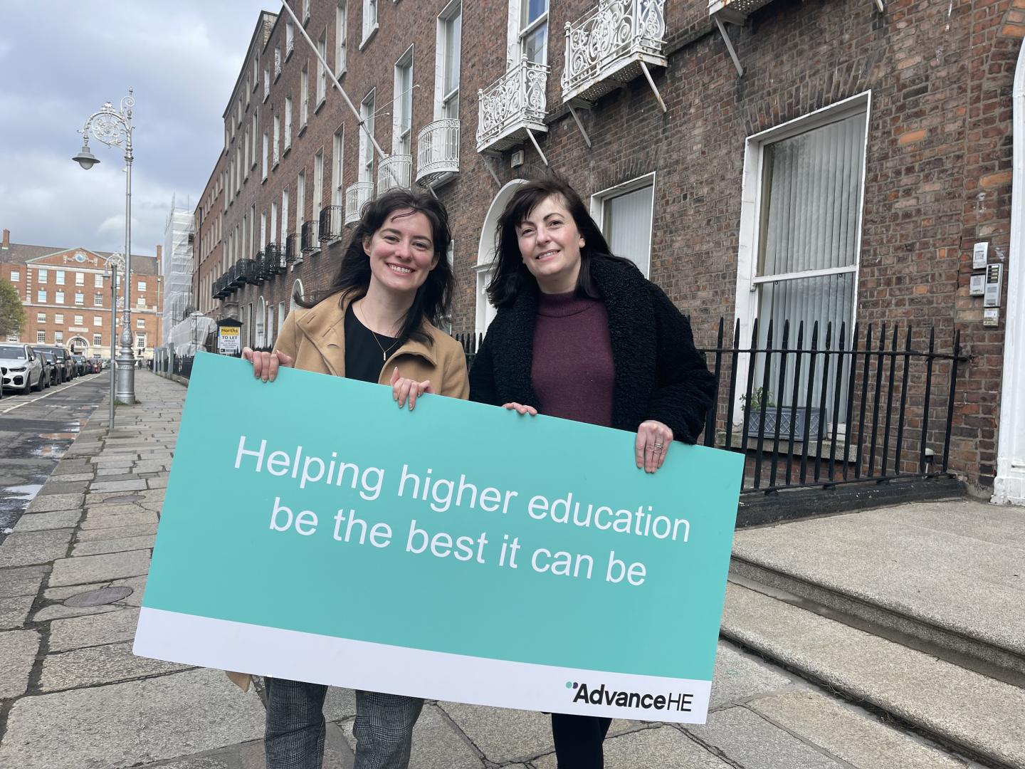 Two women holding a sign which has the Advance HE logo and says 'Helping Higher Education to be the best it can be'