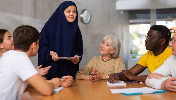 Friendly interested young Asian female student in traditional muslim black dress and khimar in discussion with group of various ages and nationalities sitting around table