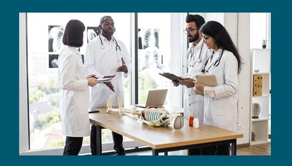 Students in lab coats listen to a medical educator with a model skeleton laid out on the table in front of them