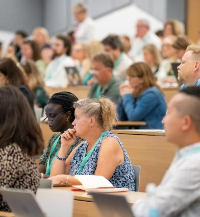 A large lecture hall filled with attendees seated at long desks, listening to a presentation. Many people are facing forward toward the front of the room, where someone is standing and speaking. 