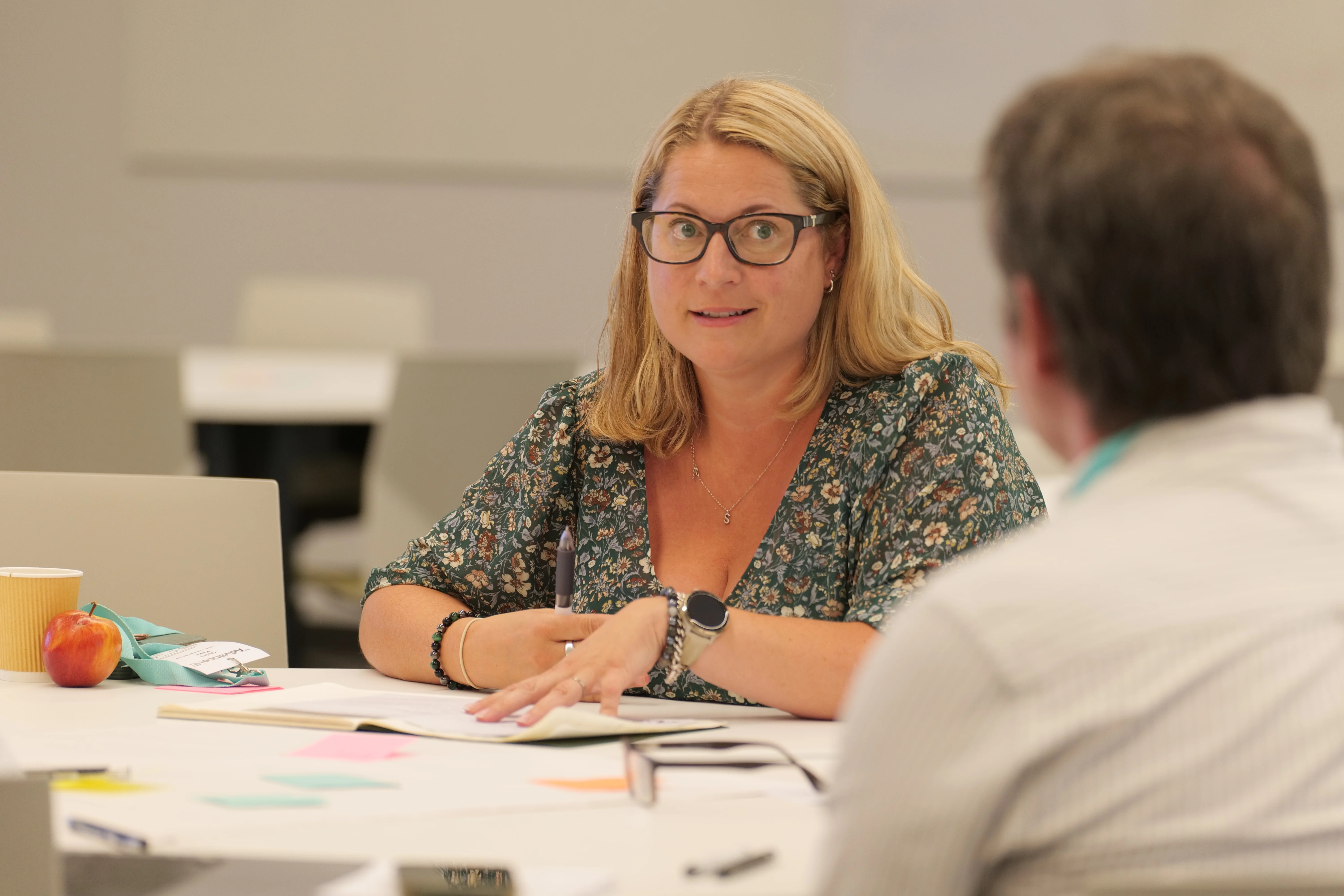 Woman at table looking through papers and talking to a colleague